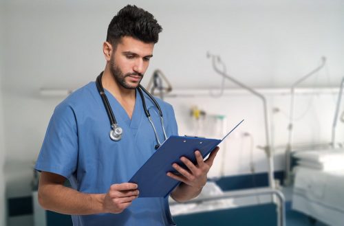 Male nurse practitioner in blue coat with stethoscope around the neck reading patient's records