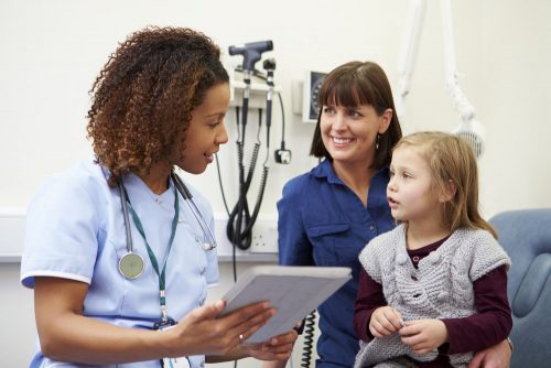 A nurse, holding a chart, talks to a child and her mother in an exam room