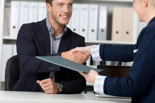 A health care lobbyist holding documents in a folder shakes hands with a client across a desk.