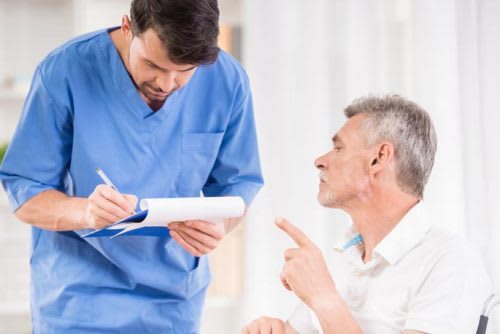 A male nurse in a blue coat writing down information provided by the elderly grey hair patient