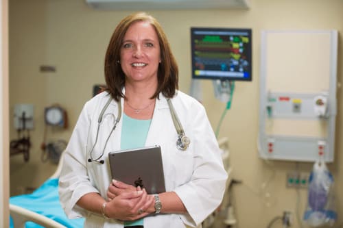 A smiling nurse practitioner holding a tablet in a hospital room.