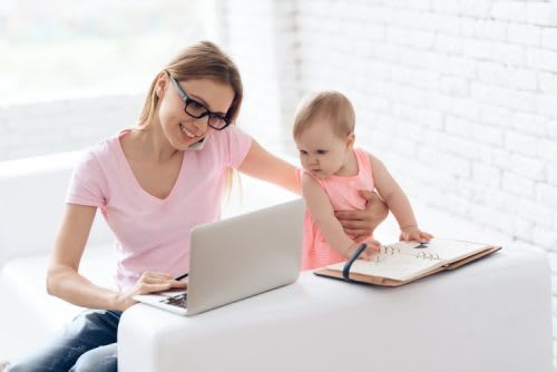 A remote employee works on a laptop while holding a young toddler.