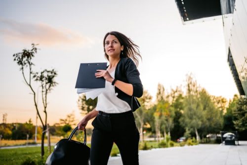 A late-to-work businessperson holding a bag and a folder of papers runs toward their office.