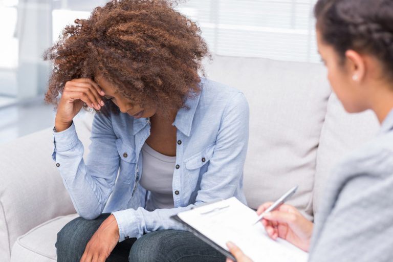 Female Doctor Taking Notes in Front of the Curly Haired Patient Holding Her Head