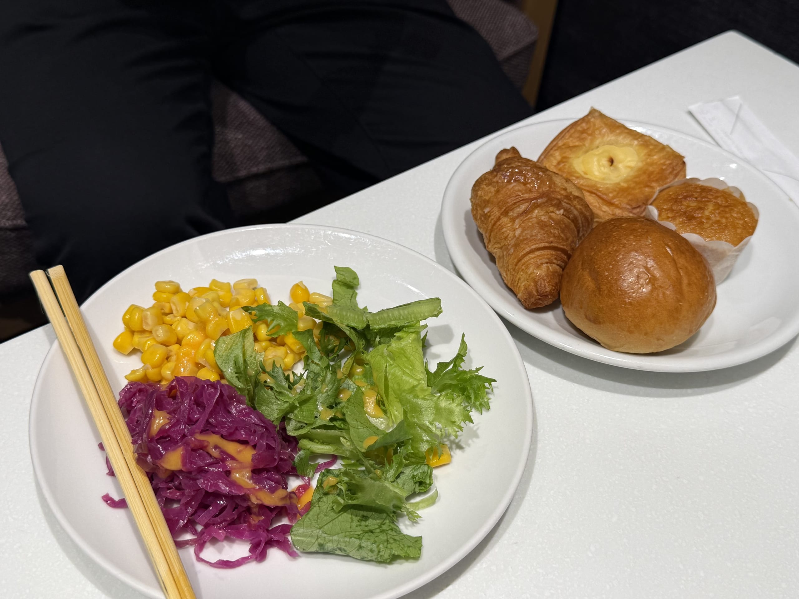 白いテーブル上のサラダ、4種のパン、割り箸の朝食。