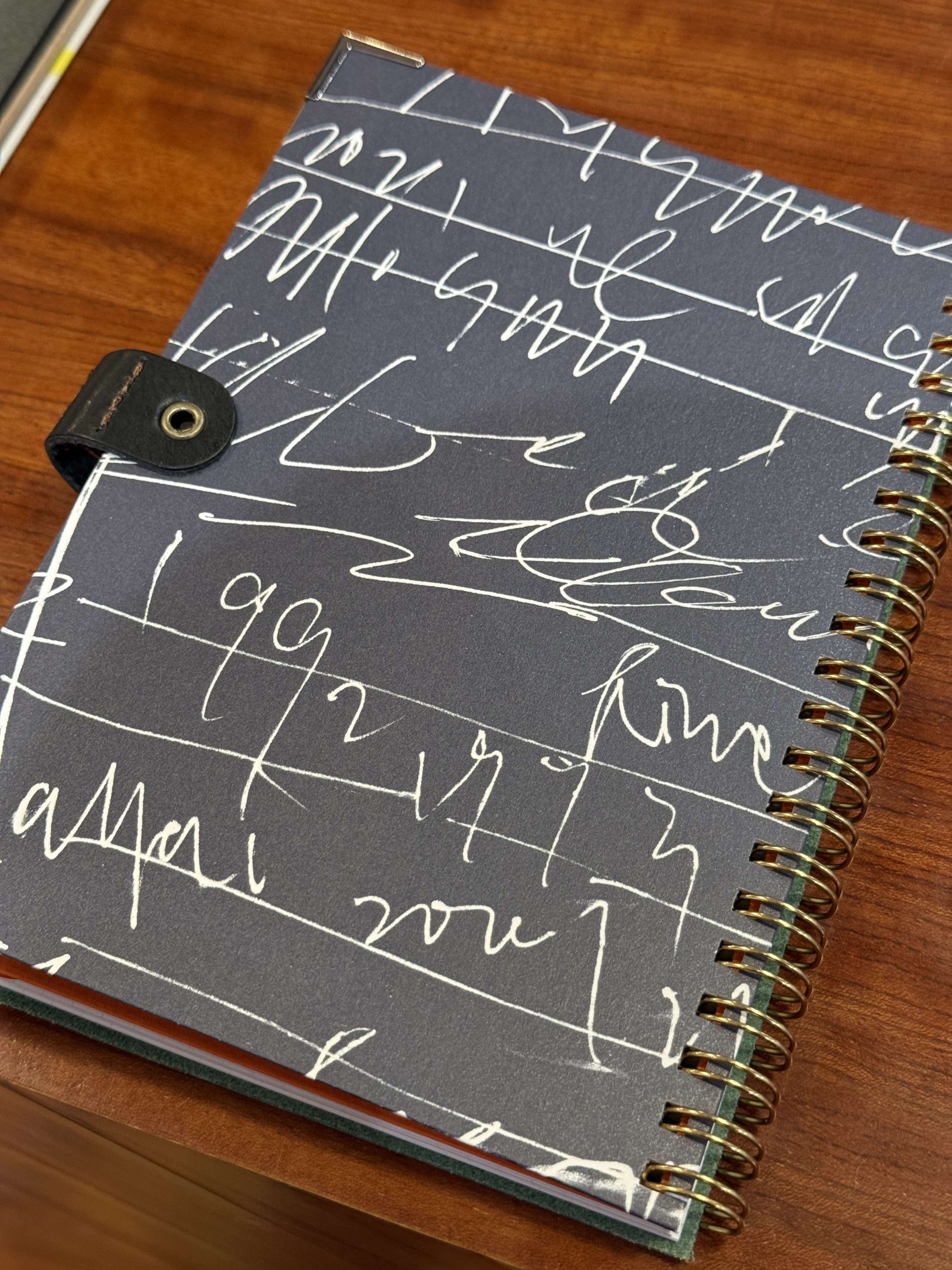 木目の机の上に置かれた、黒地に白い手書き風の線が描かれたリングノート。