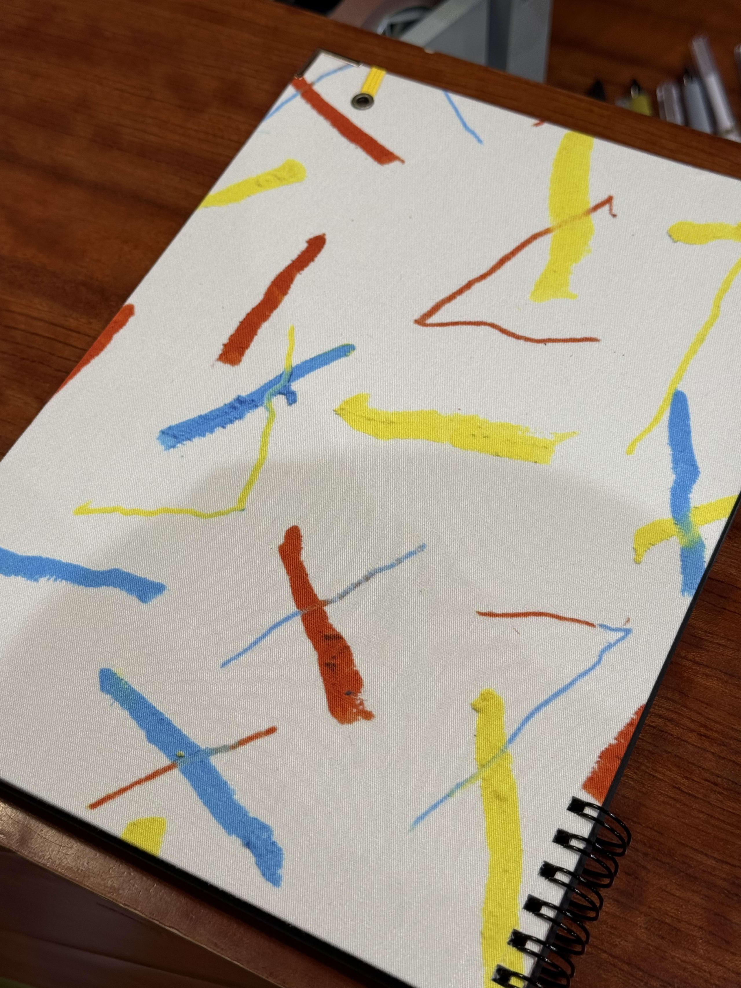 木目の机に置かれた、赤青黄の抽象模様のリングノート。