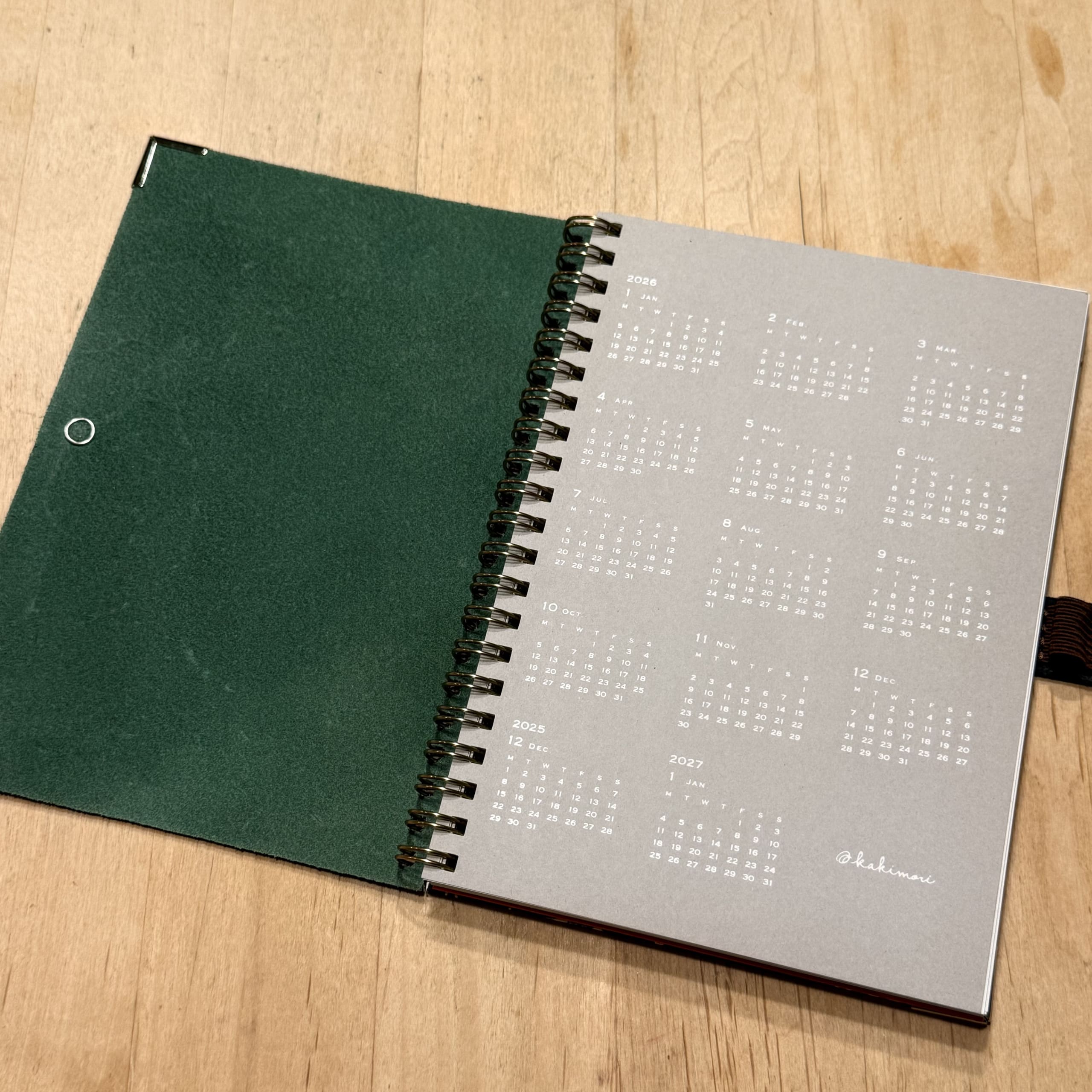 木目の机に開かれたリングノート。緑のカバーと2026年のカレンダーが見える。