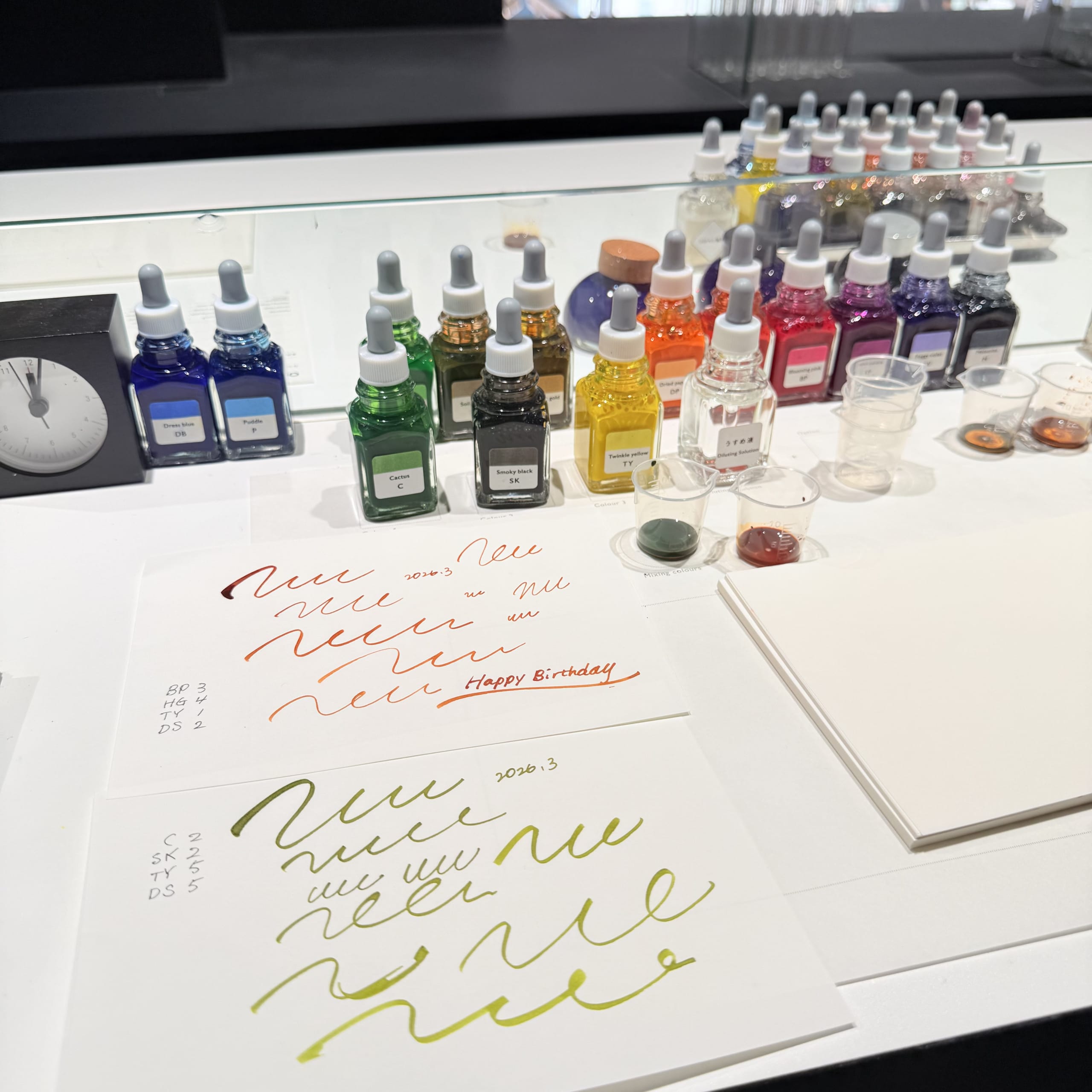 インク小瓶とビーカー、配合レシピと試し書きがされた紙が置かれたテーブル。