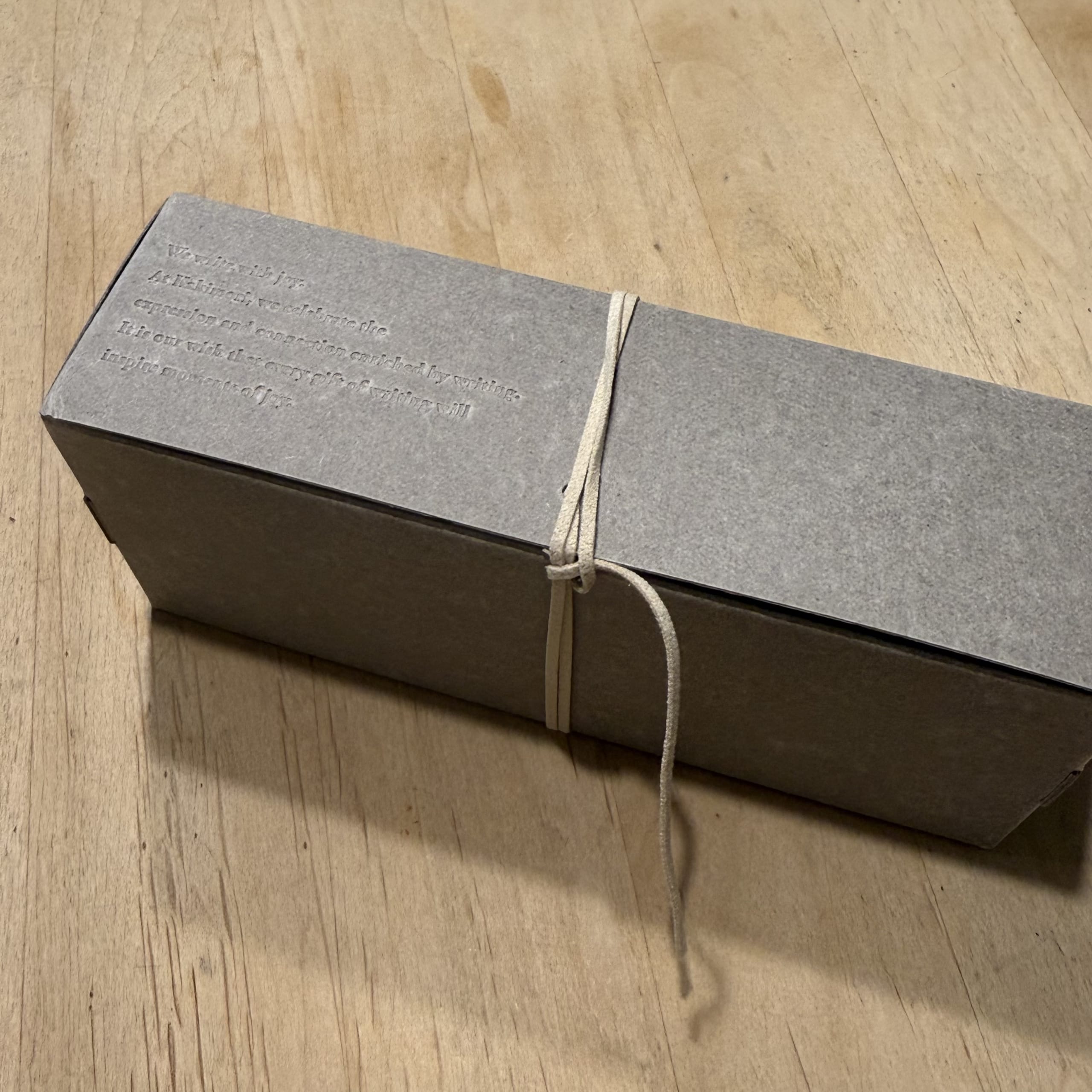 木の机に置かれたグレーの箱。エンボス文字と細い紐が特徴。