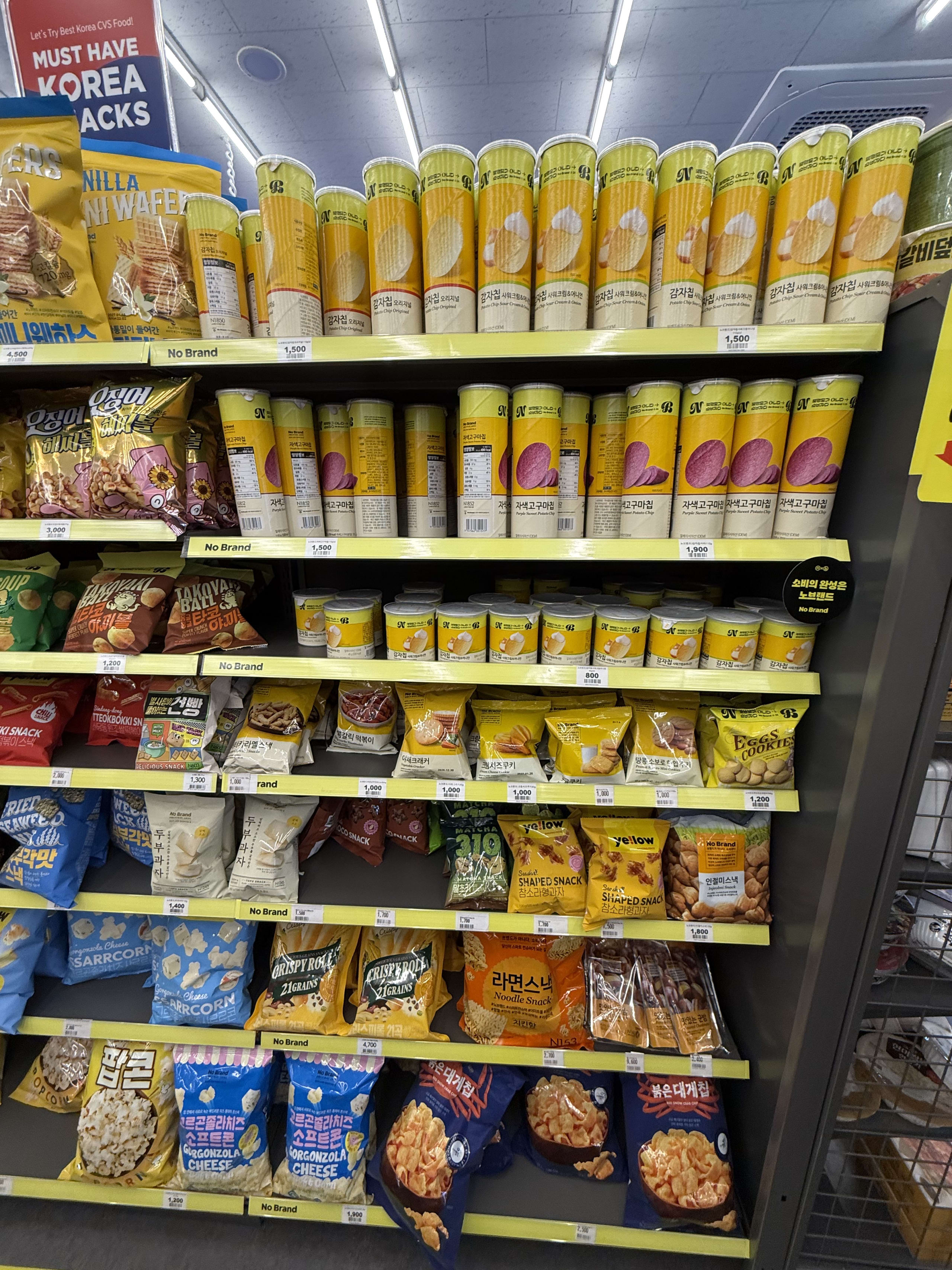 韓国のコンビニの棚に並ぶ、No Brandの黄色い筒状ポテトチップスや各種スナック菓子。