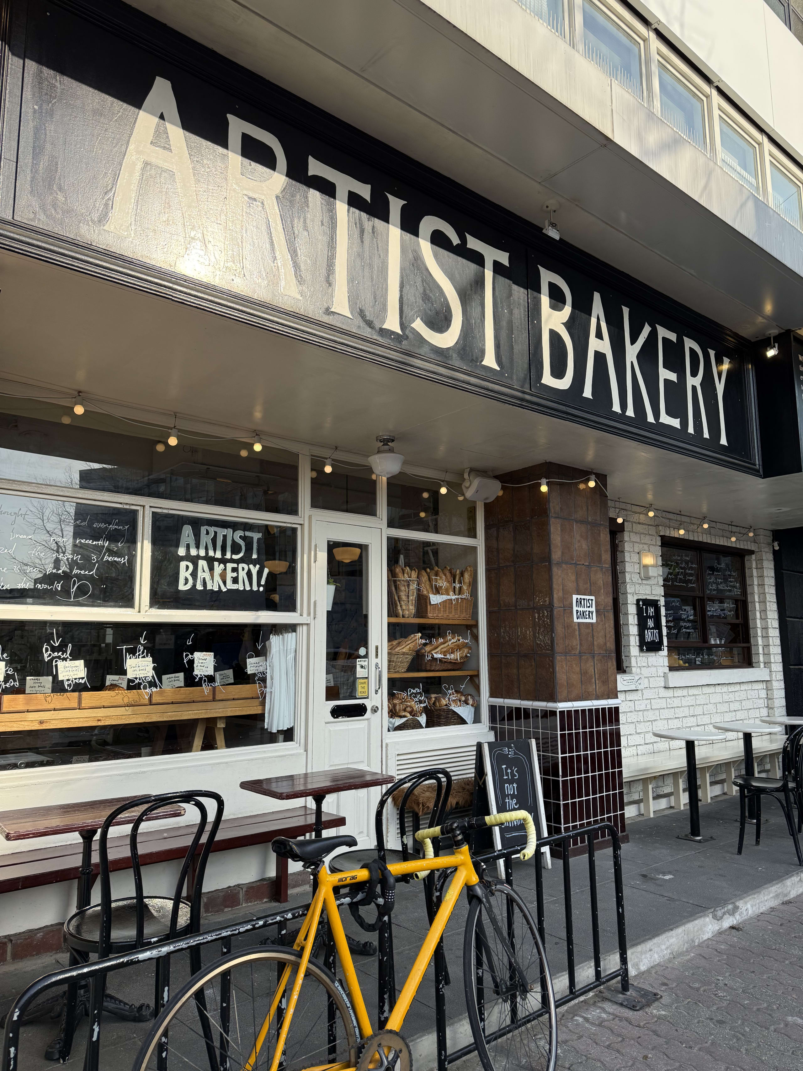 「ARTIST BAKERY」の黒い看板と店舗の外観。手前には黄色い自転車が停められている。