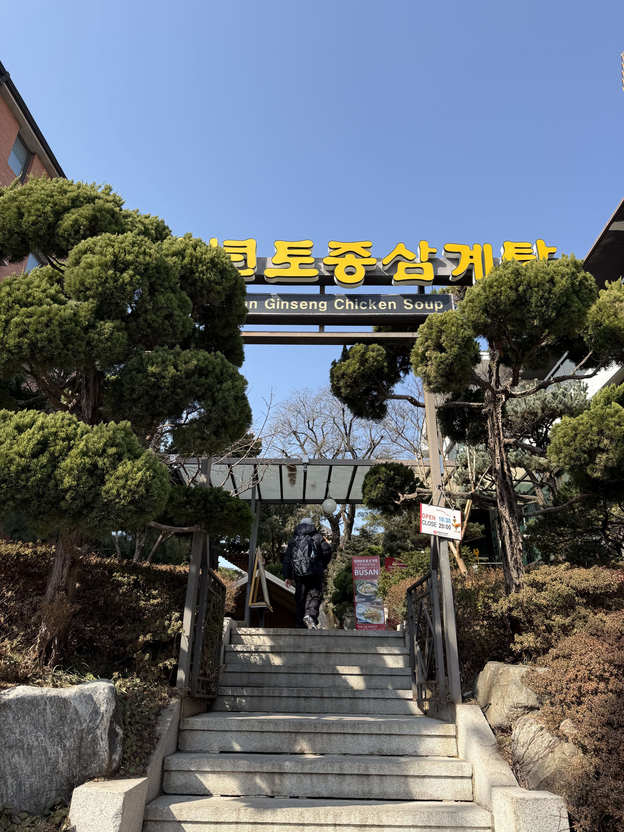 青空の下、黄色い看板が目印の参鶏湯店の入り口。石段を登る人がいる。