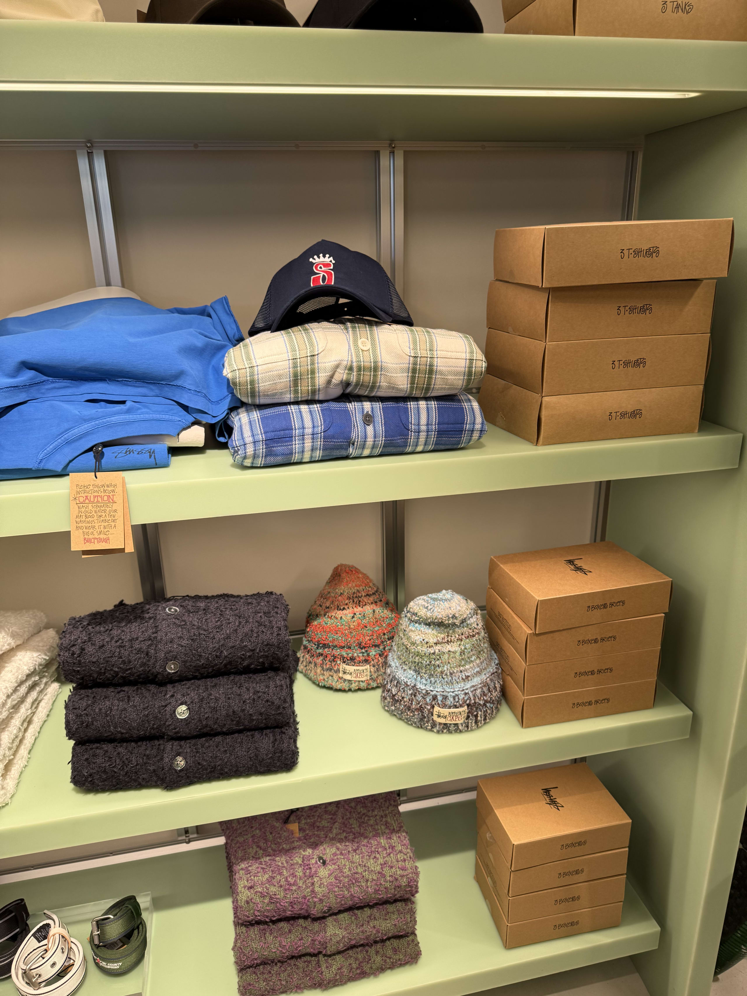 淡いグリーンの棚に並ぶステューシーのモヘアカーディガン、ニット帽、シャツ、箱入り下着。