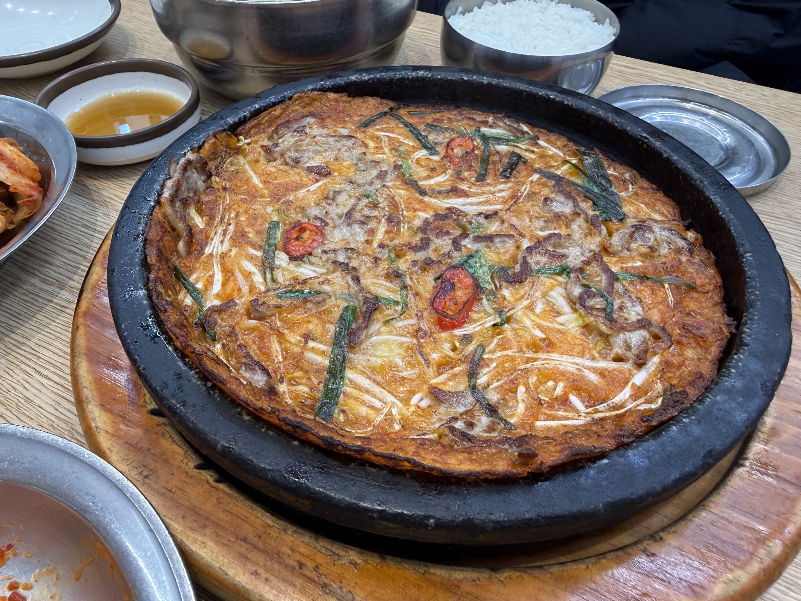 黒い鉄板に乗った大きなチヂミとキムチ、ご飯の小鉢が並ぶ。