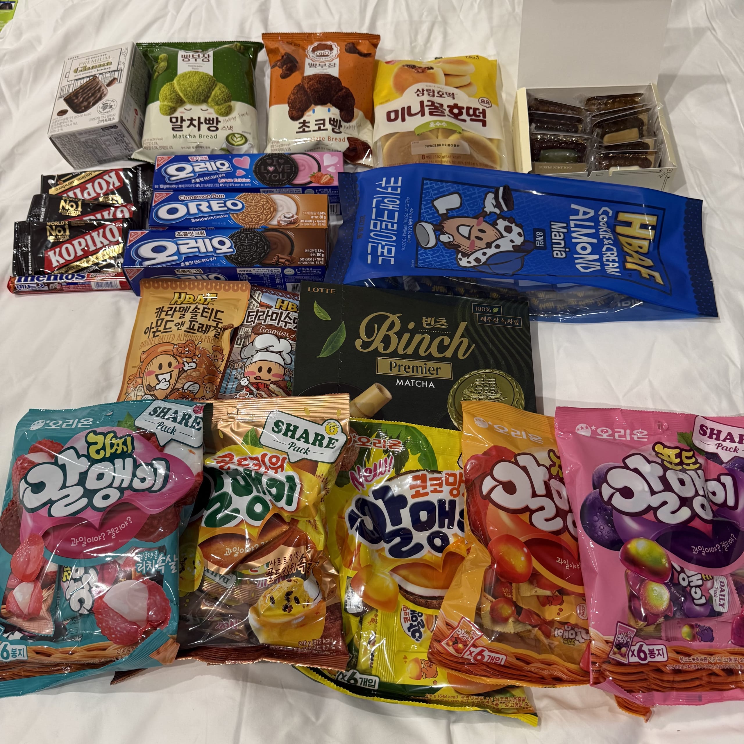 白いベッドに並べられたHBAFアーモンドやグミ等の韓国お菓子。