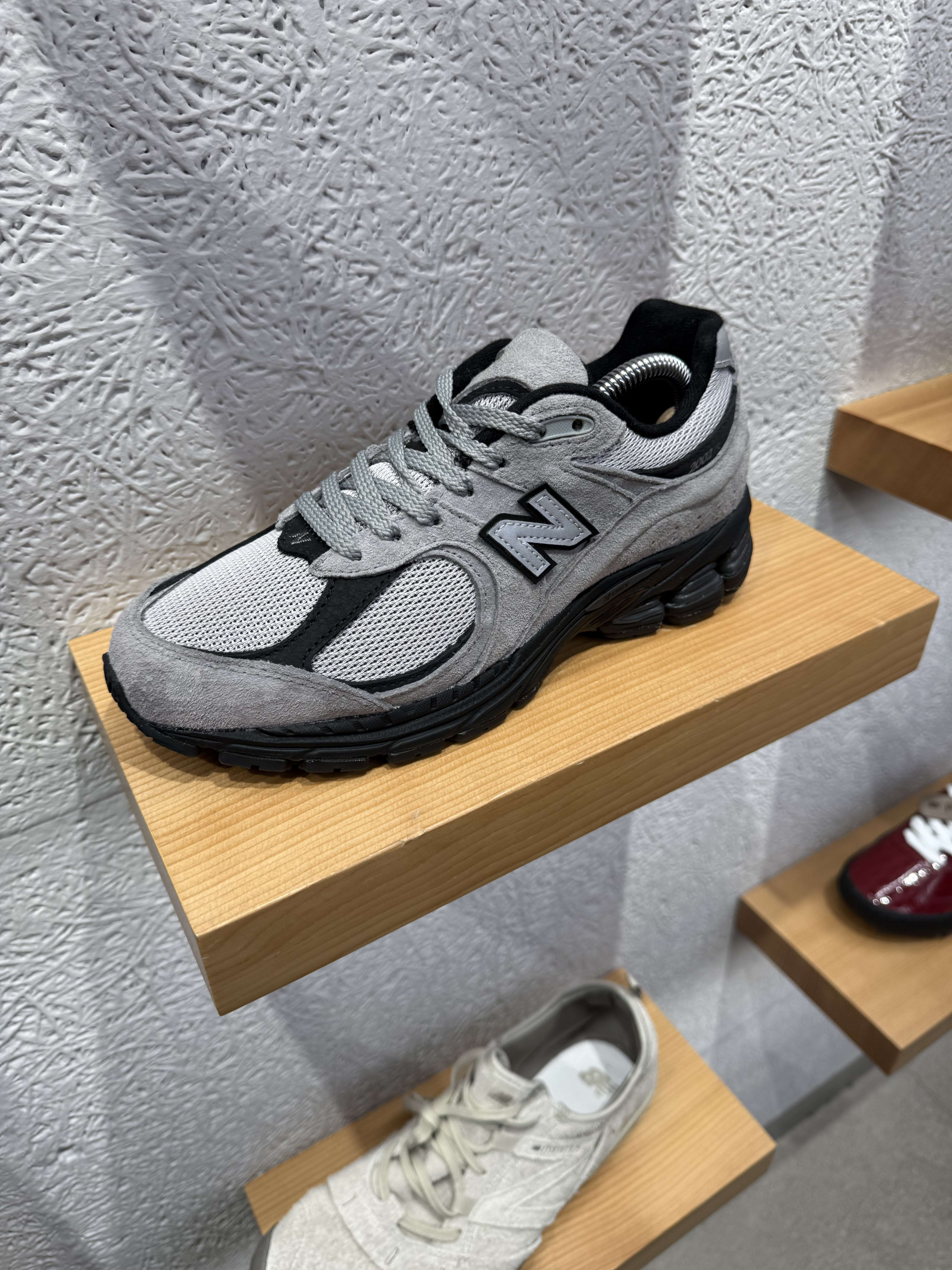 白い壁と木製棚に展示されたグレーのニューバランス製スニーカー(ミュールタイプ)。