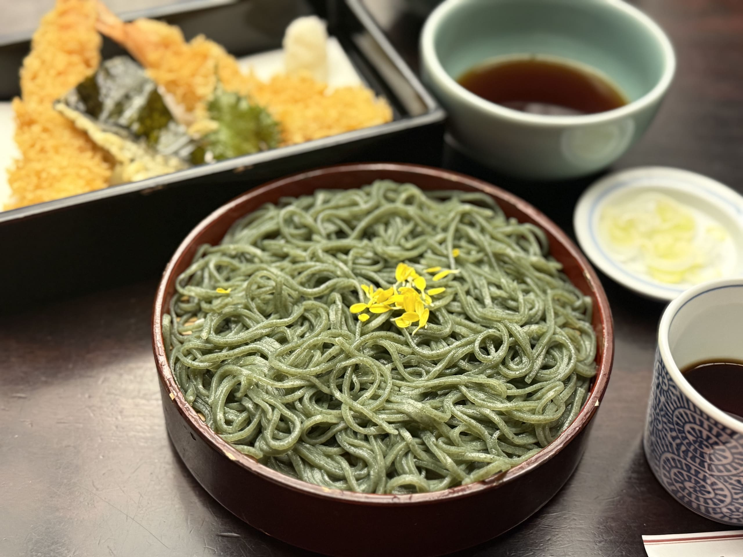 菊の花びらが乗った緑のくさきり蕎麦と天ぷら。
