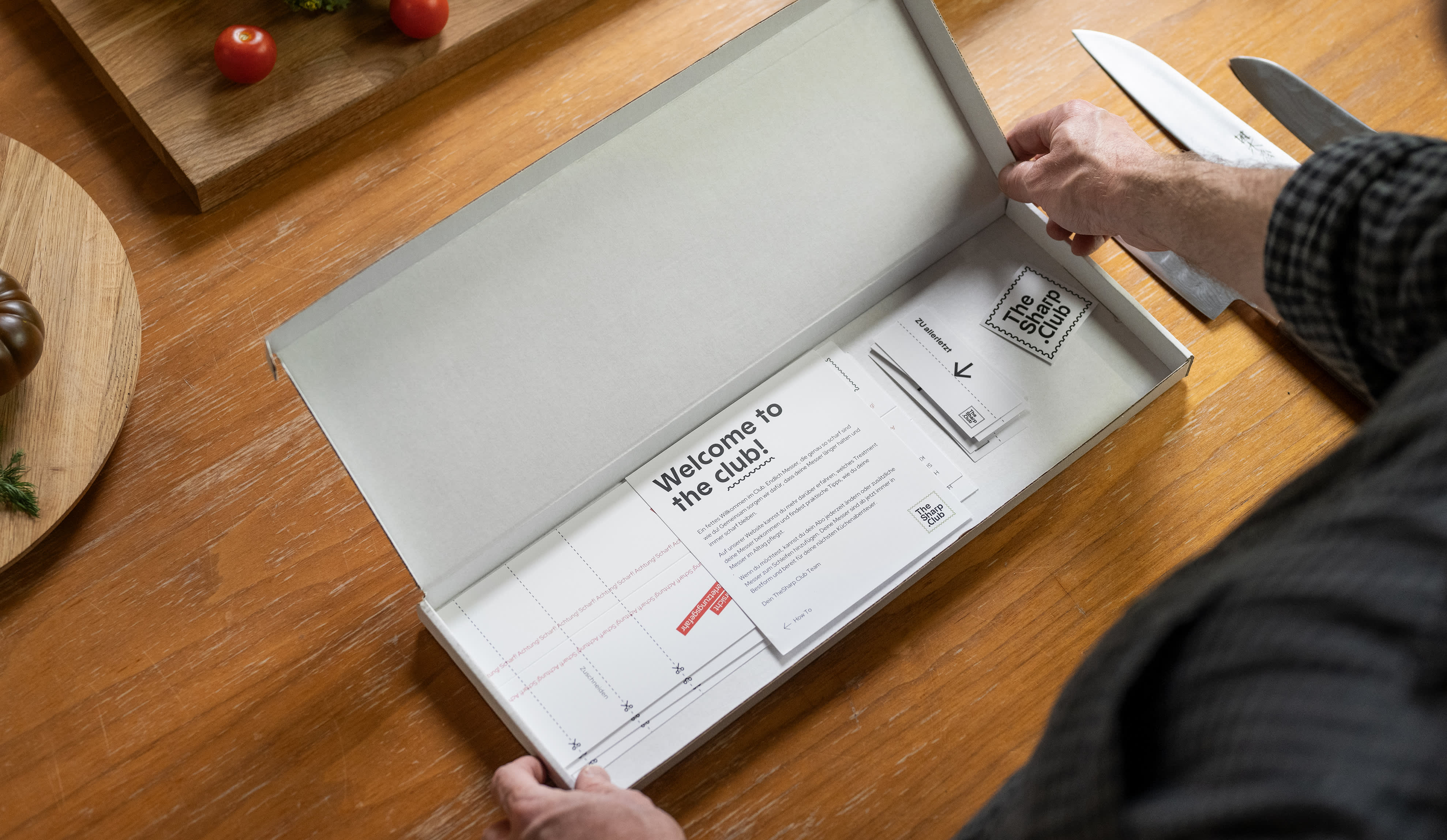 An open cardboard box lies on a wooden table between vegetables and knives. Several stickers, flyers, and other printed products from TheSharp.Club can be seen inside the box.