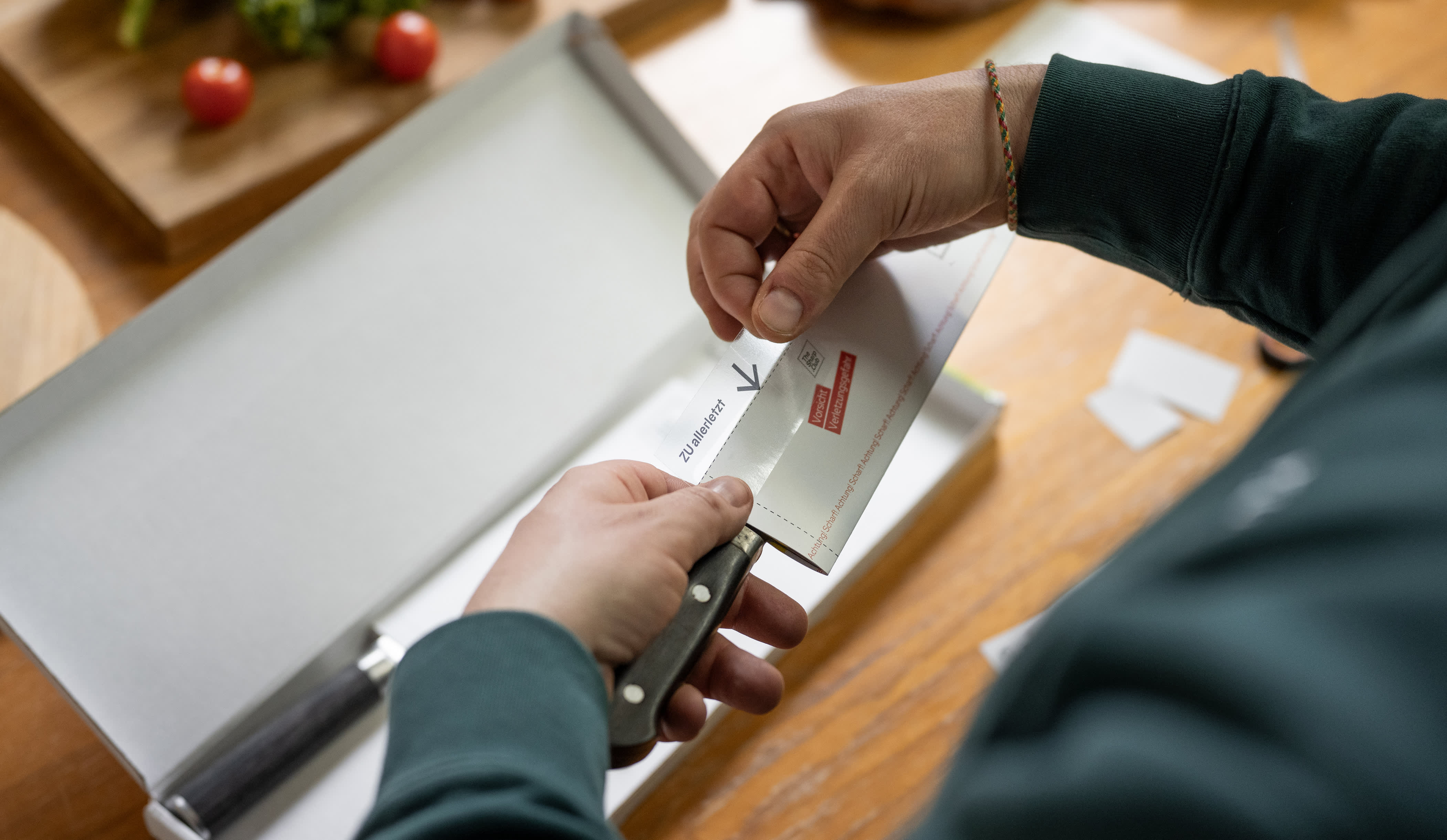 A person is using both hands to stick a transparent security sticker onto a packaged knife. Vegetables, other knives, and an open box can be seen in the background.