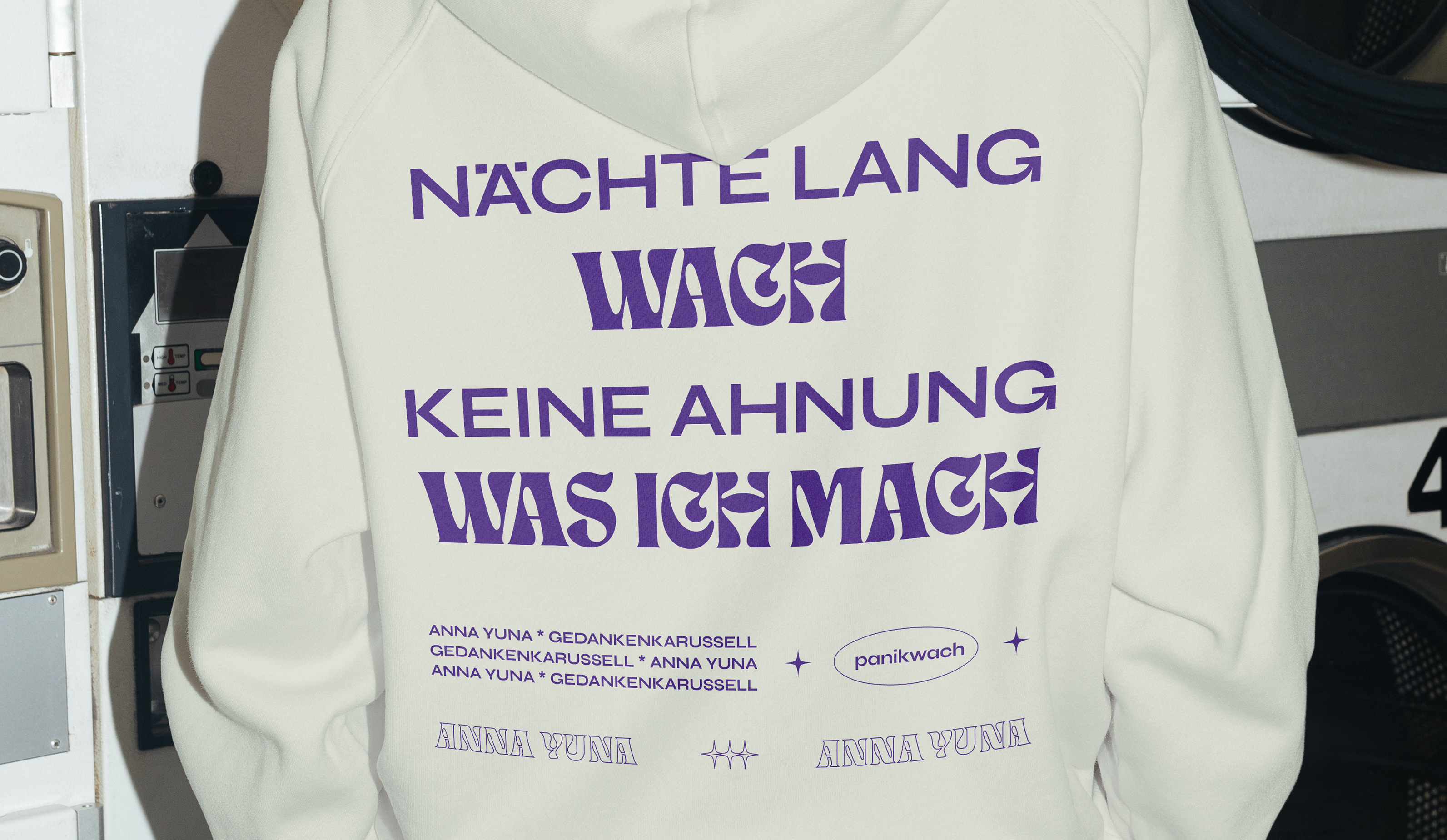 One person is standing with their back to the camera and is wearing a beige hoodie. A close-up of a purple print with different fonts of the musician ANNA YUNA can be seen on it.