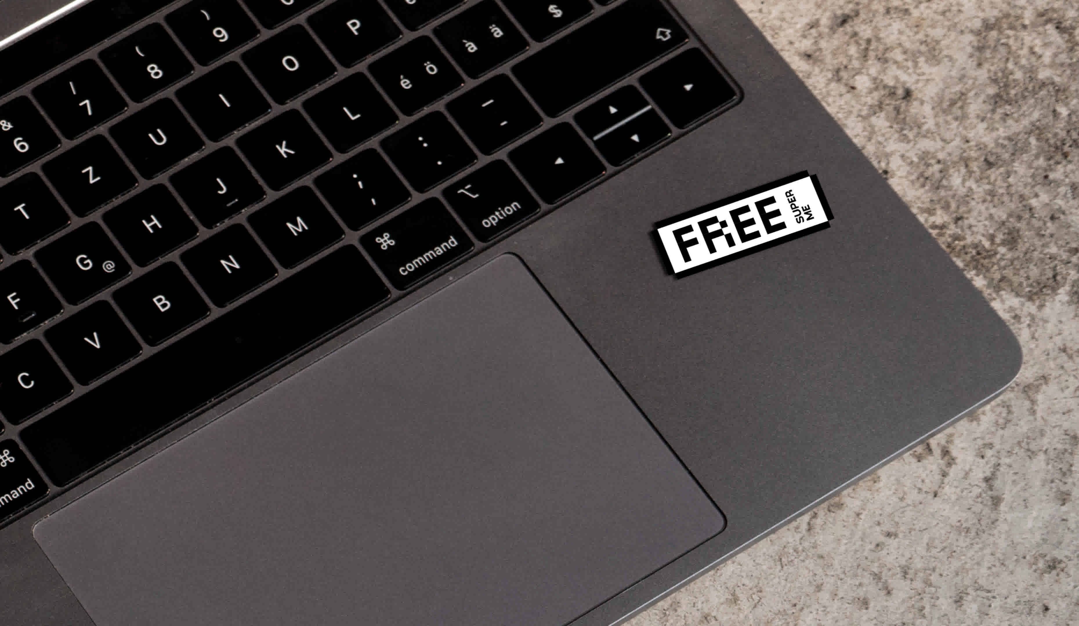 A laptop is standing on a stone floor. A sticker can be seen on the right-hand surface next to the touch pad. It shows a statement and stands out.