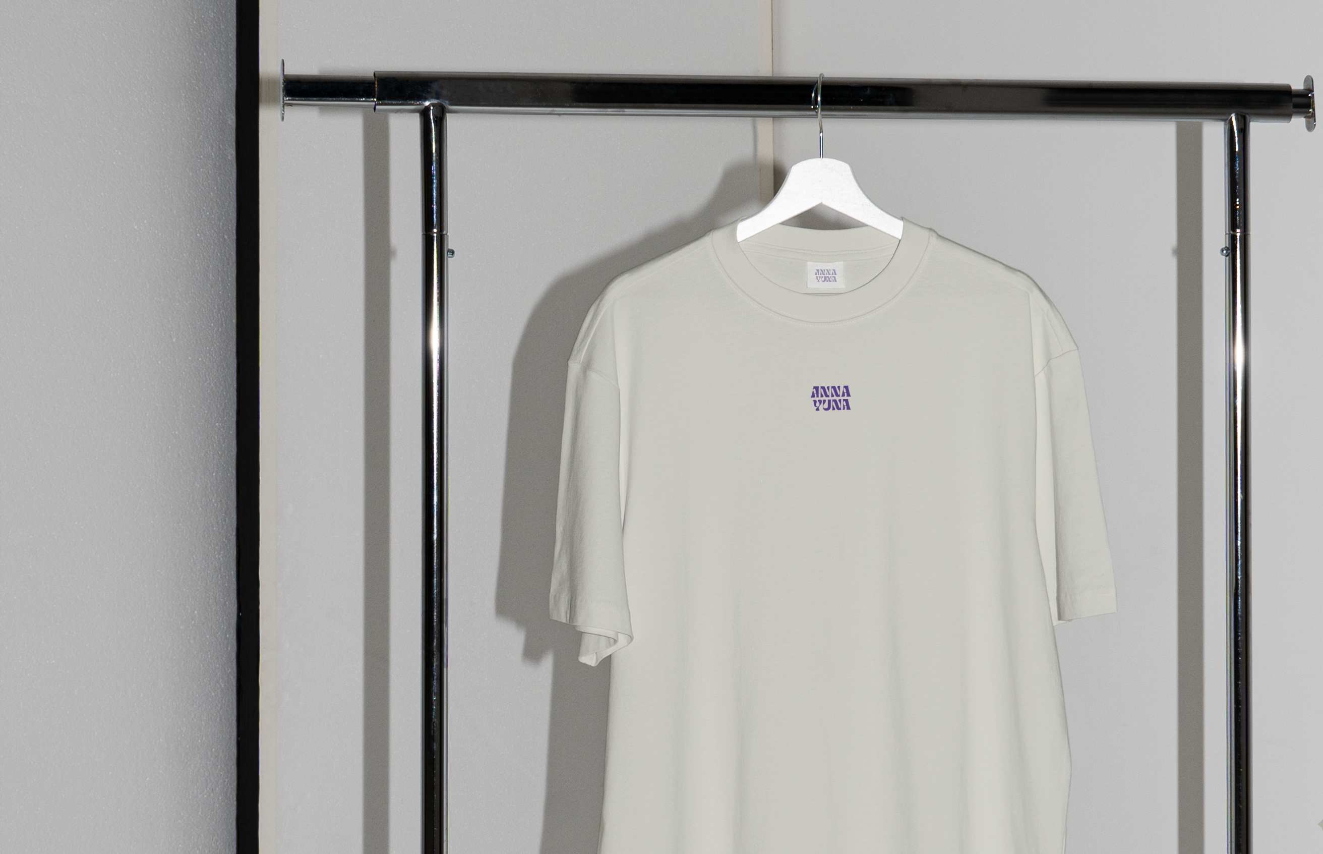 A beige shirt is hanging on a clothes rail. The shirt has a small print on the front. It says ANNA YUNA in purple.