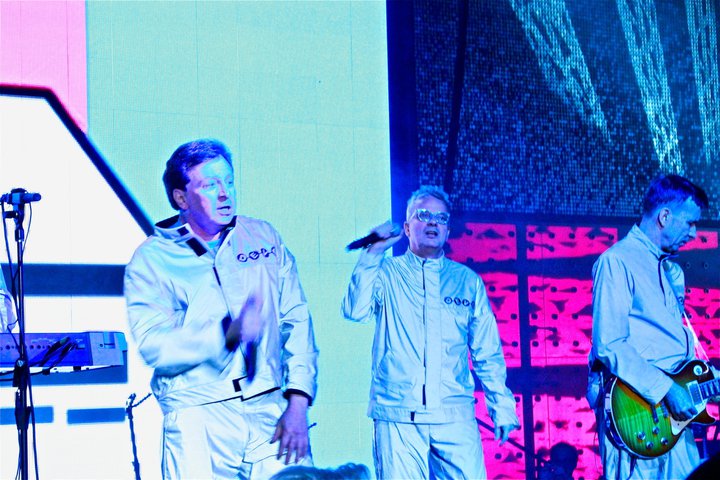Devo band performing in white jumpsuits with pink and blue stage lighting