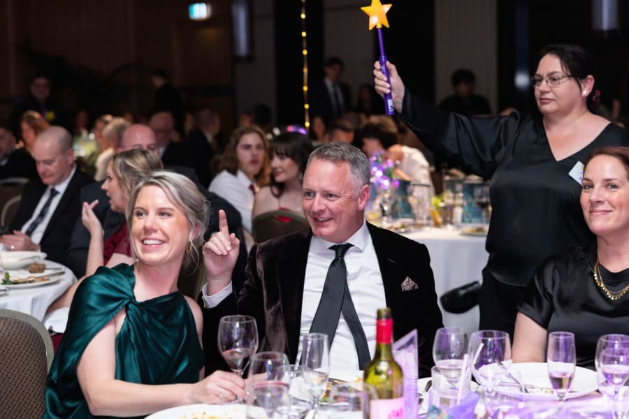 Gala guests smiling at their dinner table while a star wish wand is raised behind them