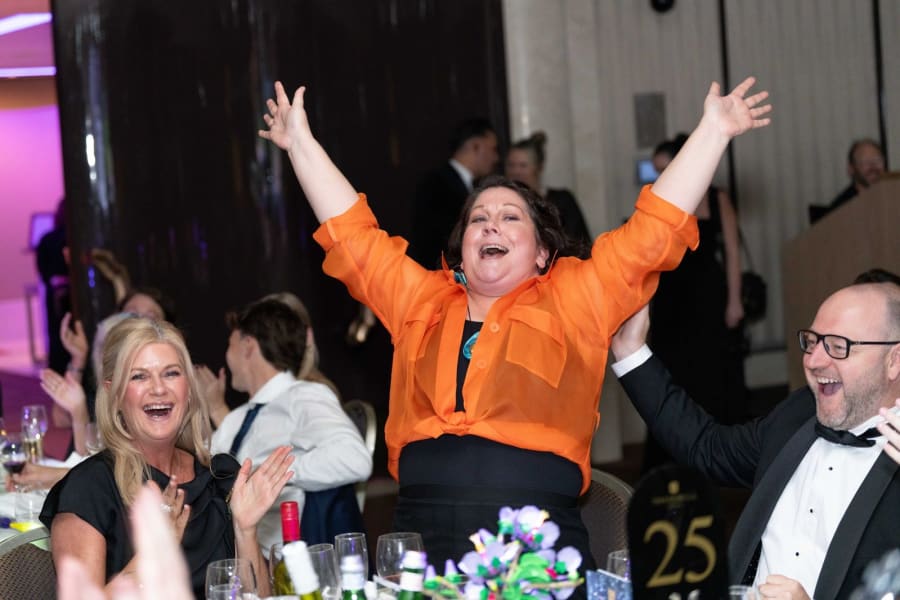 Guest in an orange shirt celebrating a winning auction bid with arms raised while her table cheers