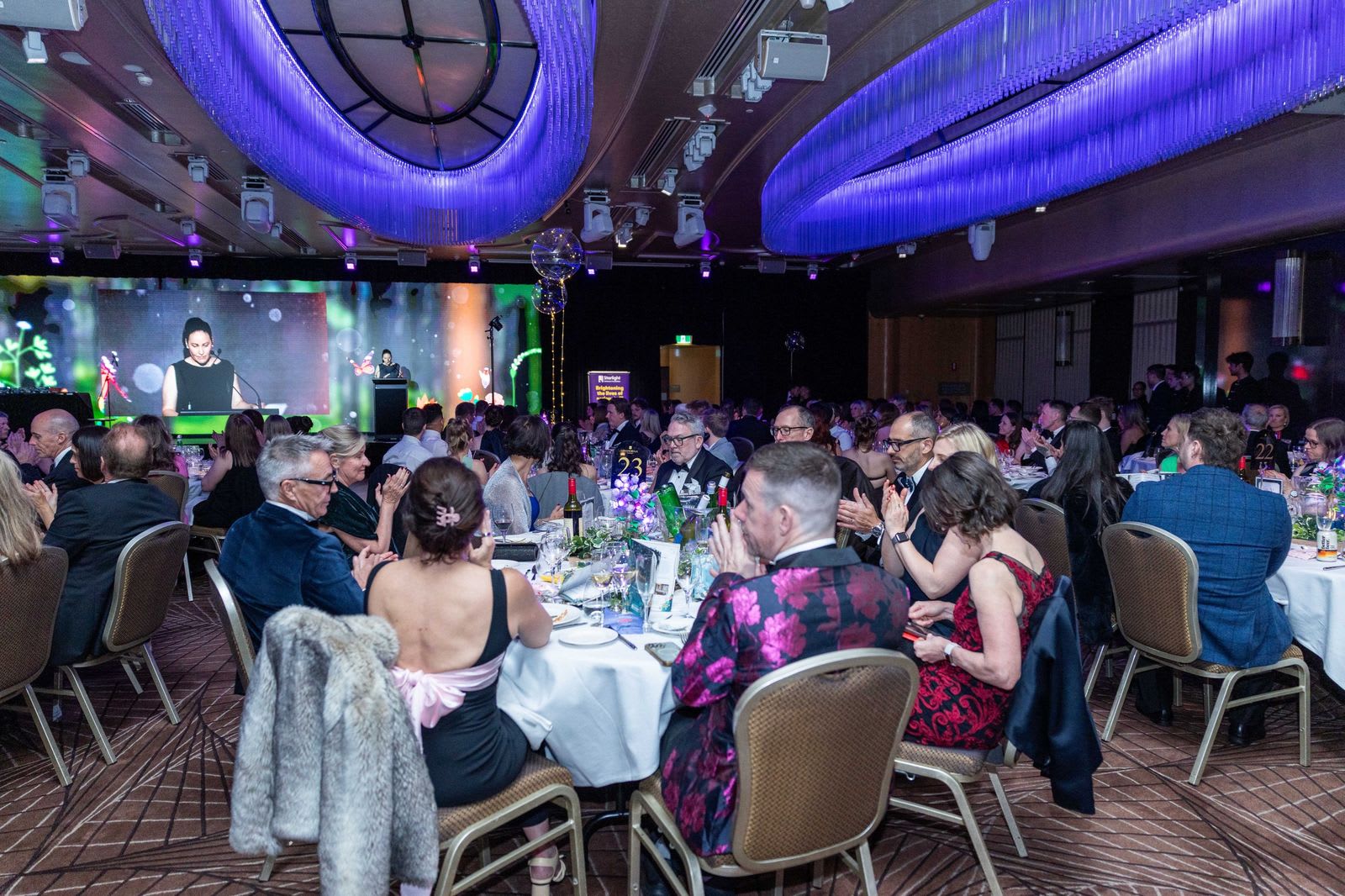 Wide view of a Sydney hotel ballroom during a gala keynote, with blue chandeliers, round dinner tables and the speaker on stage