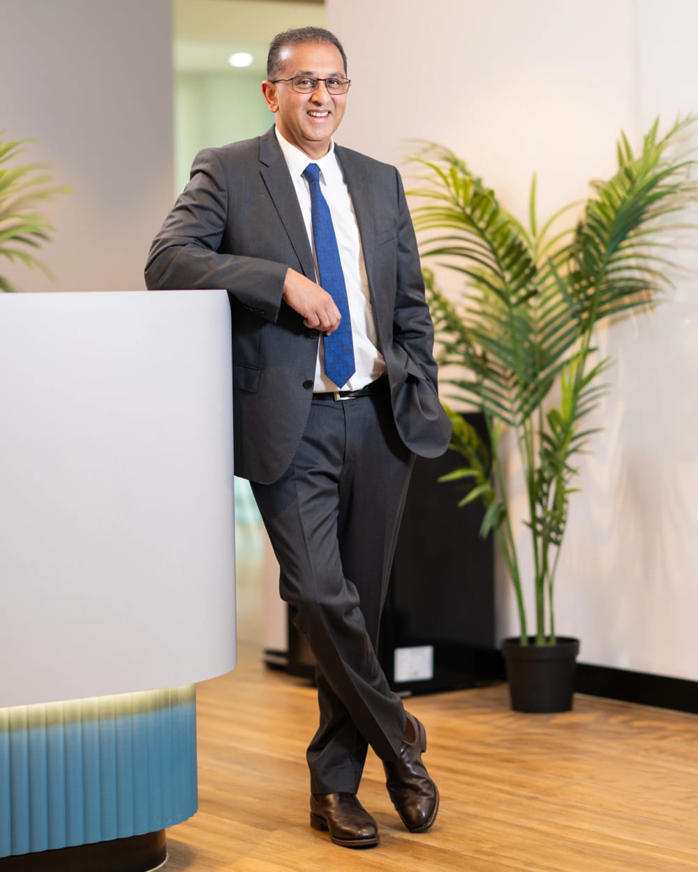 Smiling executive portrait of Dr Sris Baskaranathan in a grey suit and blue tie leaning on a reception desk at his Sydney practice