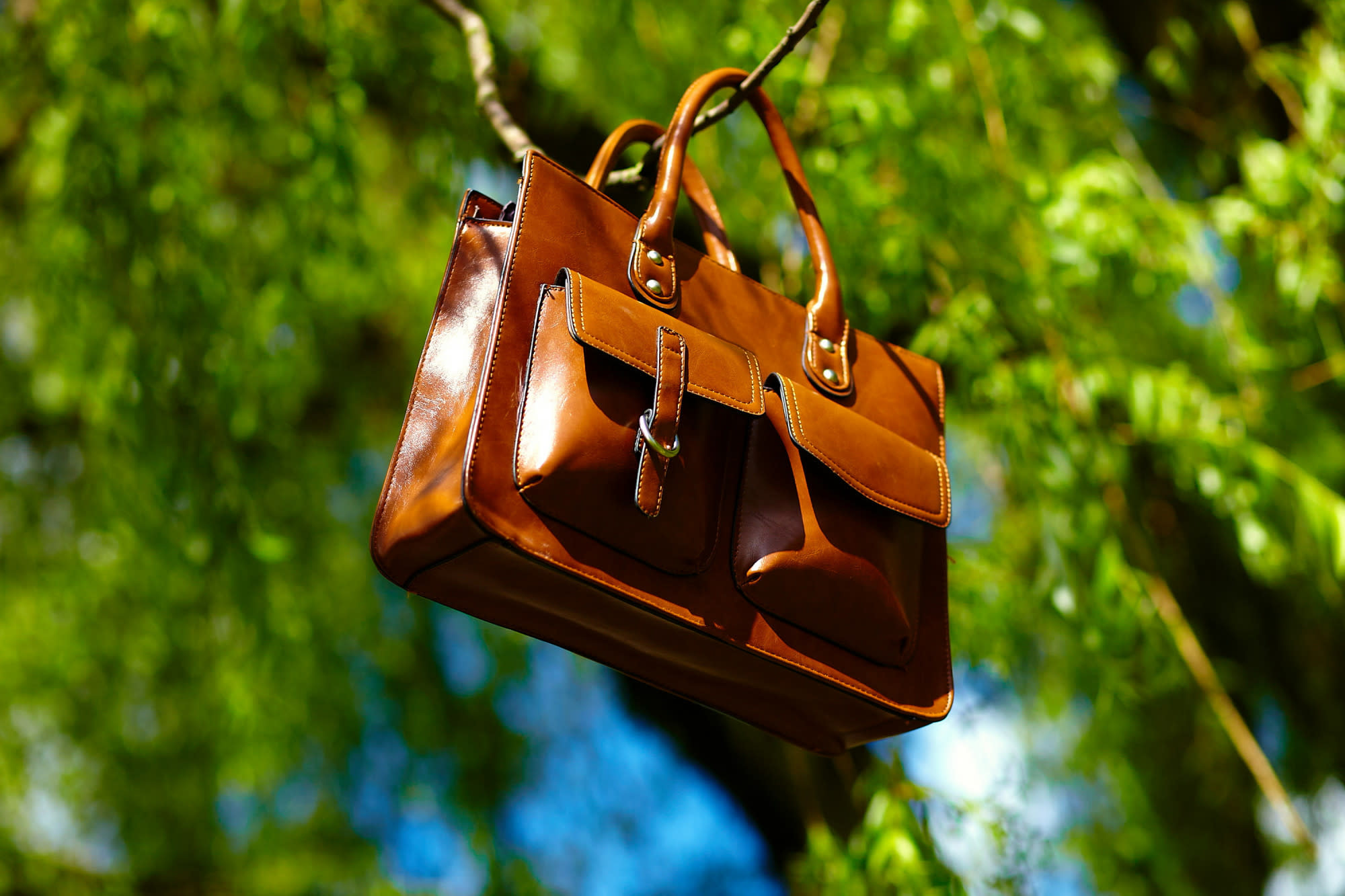 A polished brown leather satchel hanging from a tree branch against a lush green background