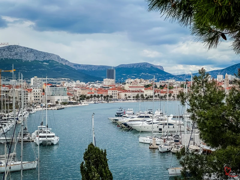 View of Split harbor with yachts and mountains in the background under a cloudy sky.
