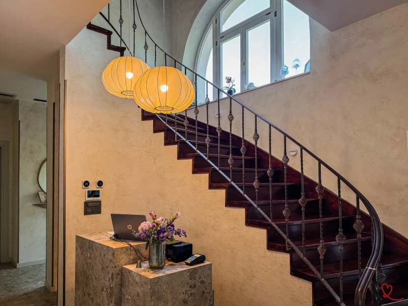 Escalier avec rambarde en fer forgé, lampes suspendues et vase de fleurs sur un meuble.