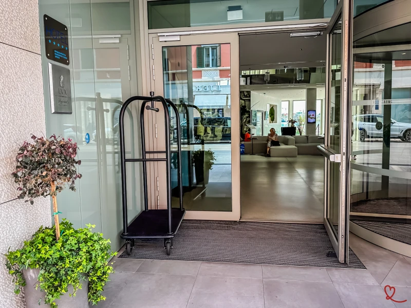 Entrada de um hotel com um carrinho de bagagem e hóspedes no átrio.