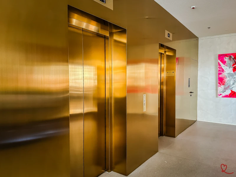Elevadores de metal dourado num átrio moderno com uma obra de arte na parede.