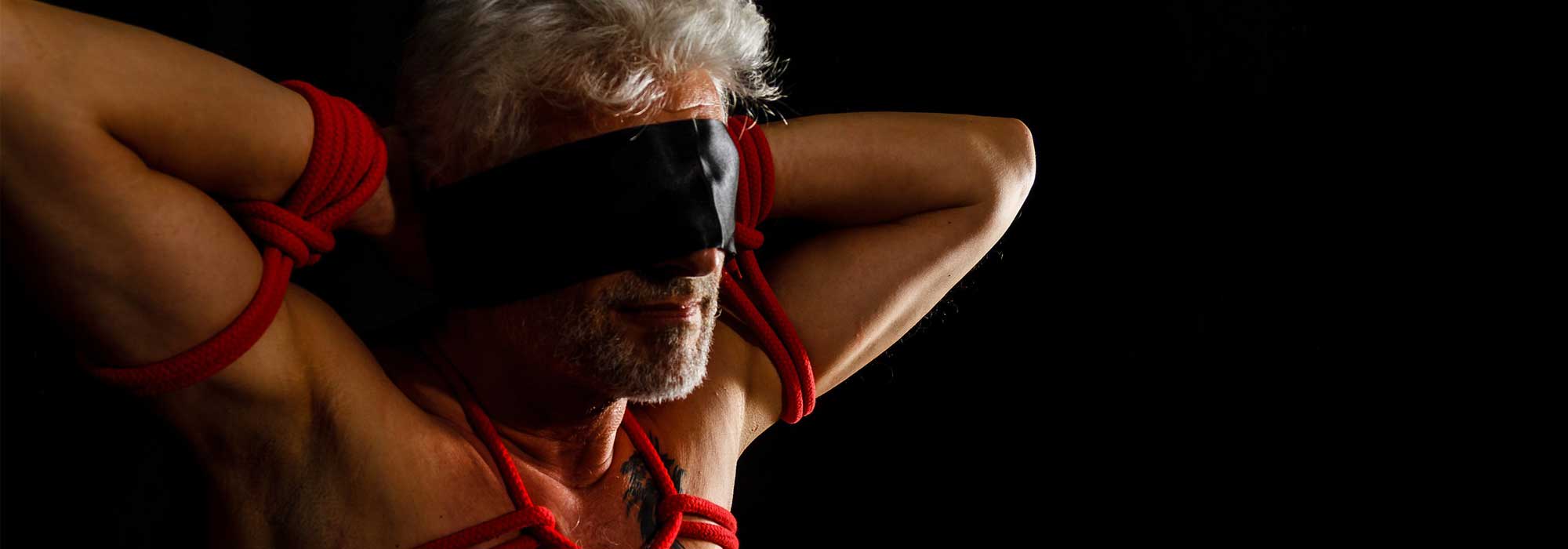 man tied up with red bondage ropes and a black blindfold, his hands are behind his head