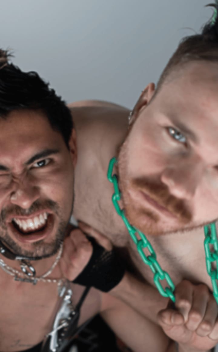 Image of two men wearing bondage gear, one has a green chain around his neck and green flecked hair, the other has a see-through top on