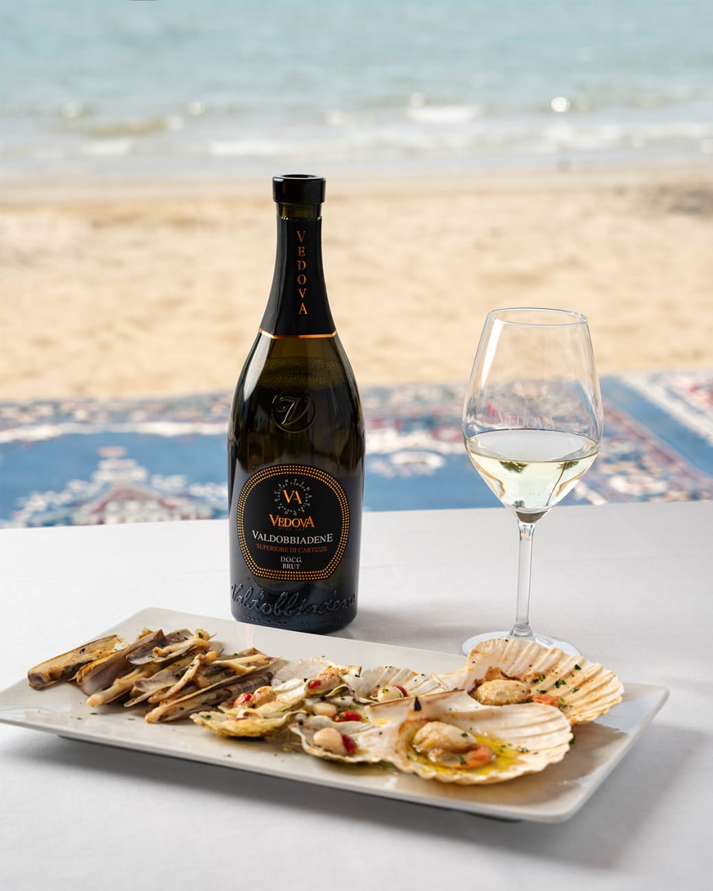 Bottiglia di Vedova Valdobbiadene Superiore di Cartizze Brut e piatto di crudi e capesante su un tavolo in riva al mare