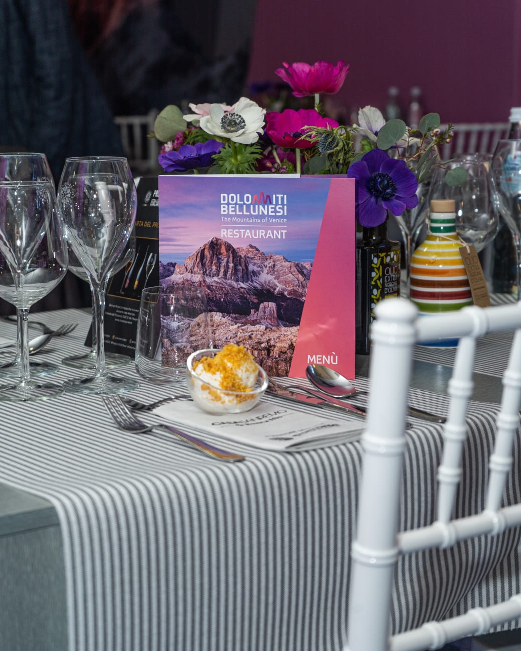 Tavolo del ristorante Dolomiti Bellunesi apparecchiato: il menù «The Mountains of Venice — Restaurant», i calici e una composizione di anemoni fucsia e viola