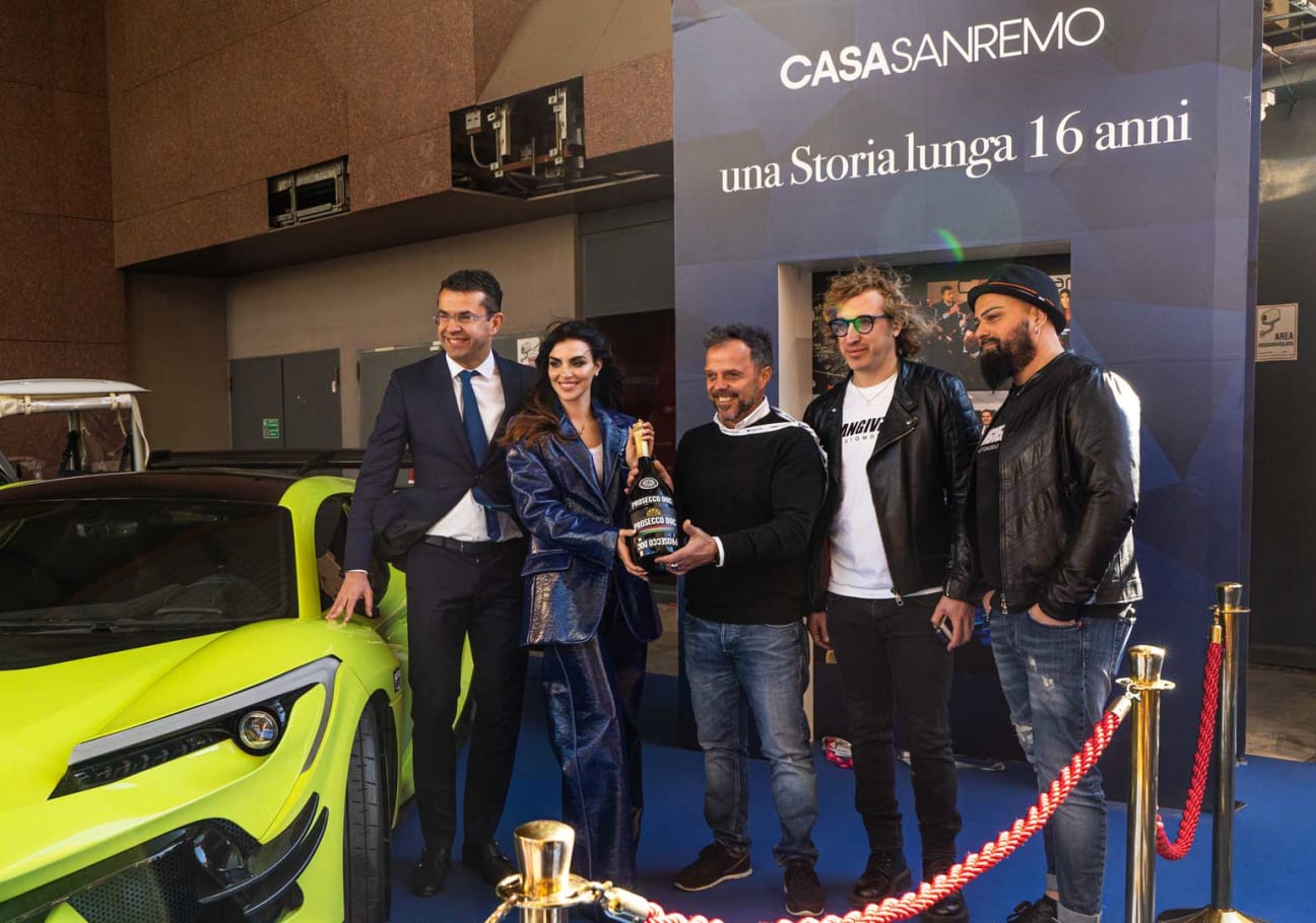Consegna di una bottiglia celebrativa accanto all’auto sportiva esposta, sotto il pannello «Casa Sanremo — una storia lunga 16 anni»