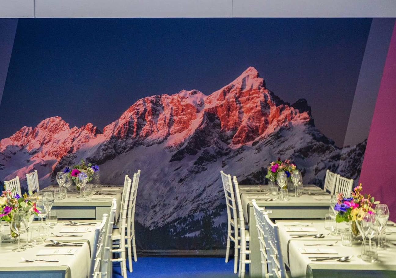 La sala ristorante con i tavoli lunghi apparecchiati davanti alla grande parete fotografica delle Dolomiti al tramonto