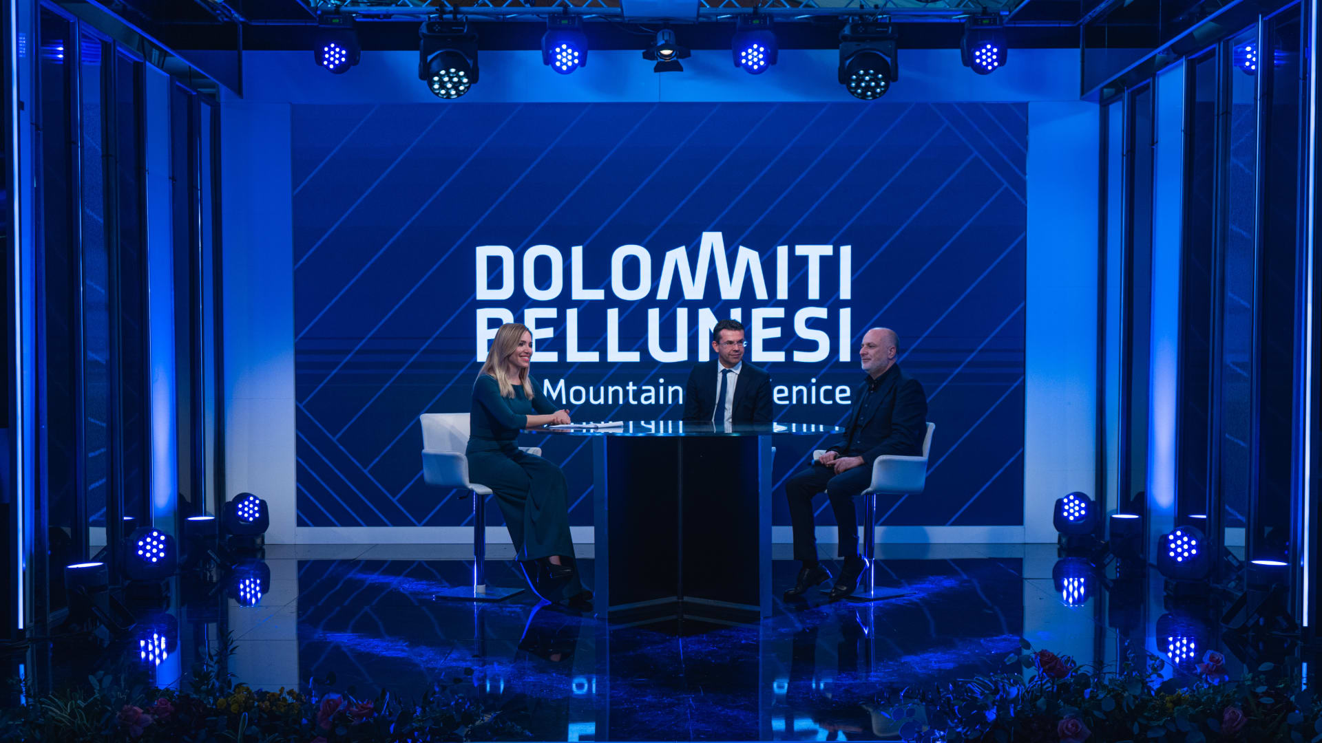 Il palco dell’area TV illuminato di blu: la conduttrice e un ospite seduti ai lati del leggio, davanti al ledwall «Dolomiti Bellunesi — The Mountains of Venice»