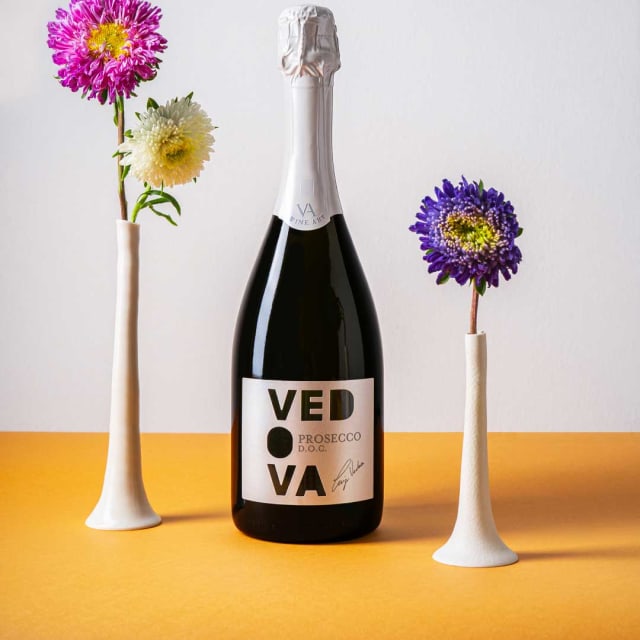Bottiglia Vedova Prosecco DOC con etichetta écru, tra due vasi bianchi con astri fucsia e viola su piano arancione