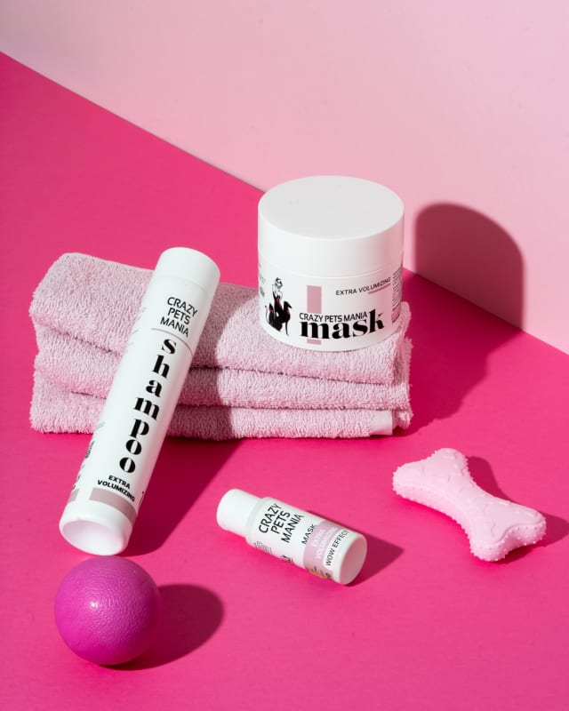 Shampoo e maschera Crazy Pets Mania appoggiati su fondo rosa e fucsia tra asciugamani piegati, un ossicino di gomma e una pallina