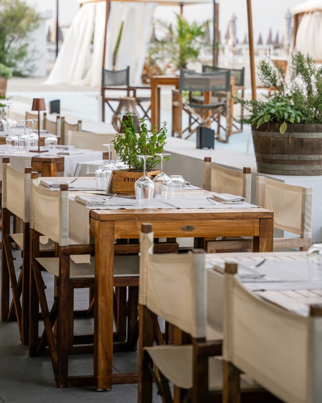 La sala del ristorante sulla sabbia apparecchiata: tavoli in teak, sedie regista beige, piantine in cassette di legno e i gazebo dalle tende bianche sullo sfondo