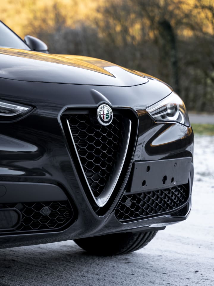 Frontale di un’Alfa Romeo Stelvio nera con lo scudetto a V, su un piazzale velato di brina