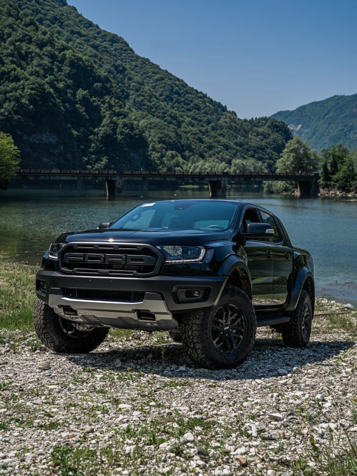 Ford Ranger Raptor nero fotografato di fronte sulla riva di un lago, con un ponte e le montagne alle spalle