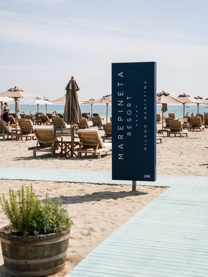 Il totem blu «Marepineta Resort · Milano Marittima · 248» piantato sulla sabbia, tra la passerella azzurra e le file di lettini e ombrelloni affacciati sul mare